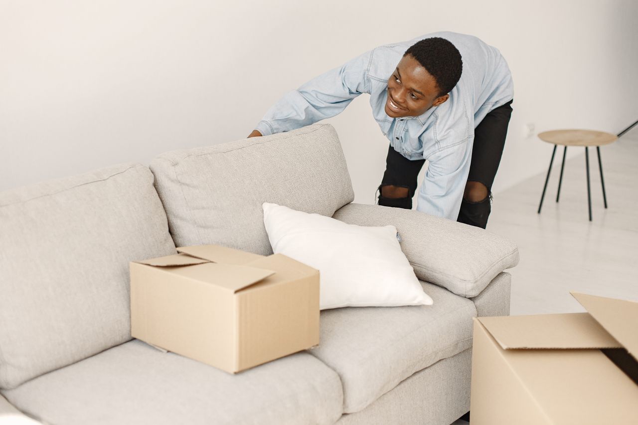 why-choose-us-03 Young man smiling while arranging cardboard boxes in a modern living room.