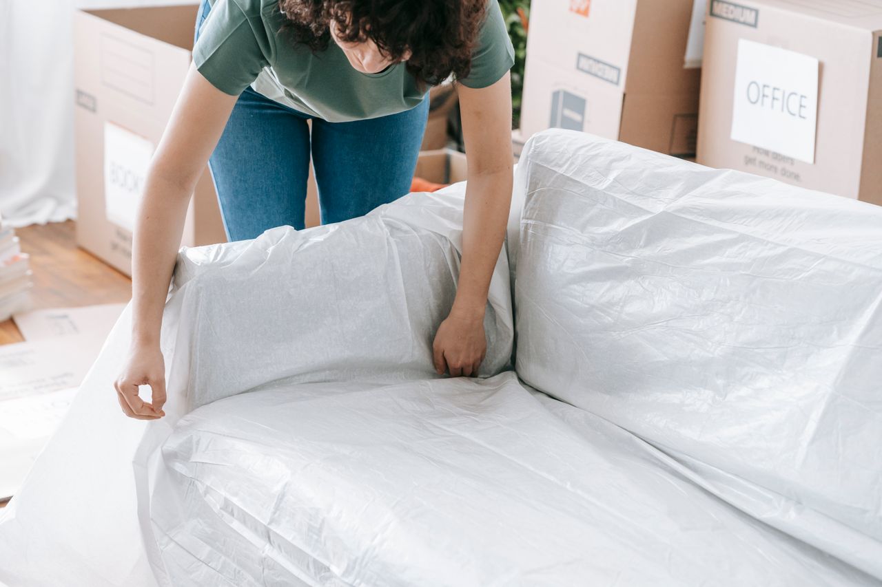 why-choose-us-02 A woman carefully wraps a sofa in protective material while packing boxes in a bright room.