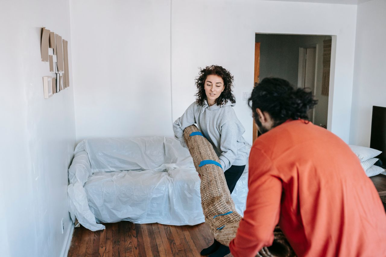 why-choose-us-03 A couple lifting a rolled carpet in a minimally furnished apartment living room.