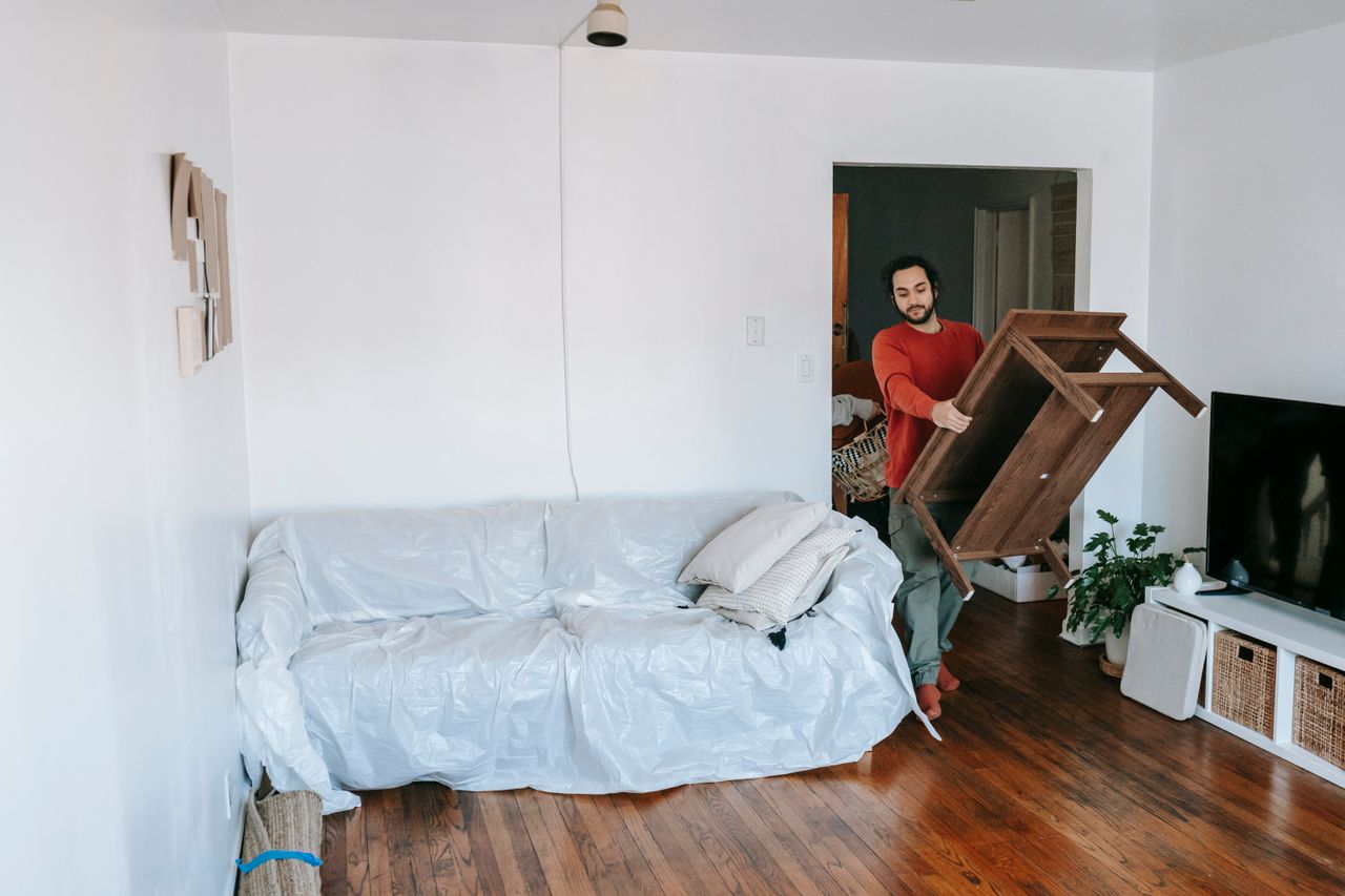 services-01 Man lifting a wooden table in a bright living room with wrapped sofa, preparing for a move.